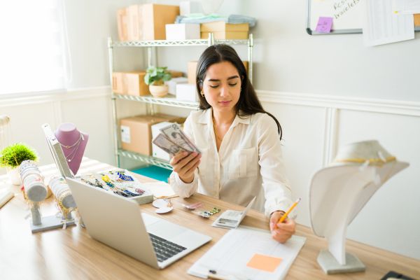A young woman sits at a desk in a brightly lit room, counting cash while surrounded by jewelry displays, a laptop, and shipping supplies.