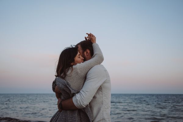 A couple hugs tightly on a beach against the backdrop of the ocean and a gradient sky. This could be a romantic moment before a marriage proposal.