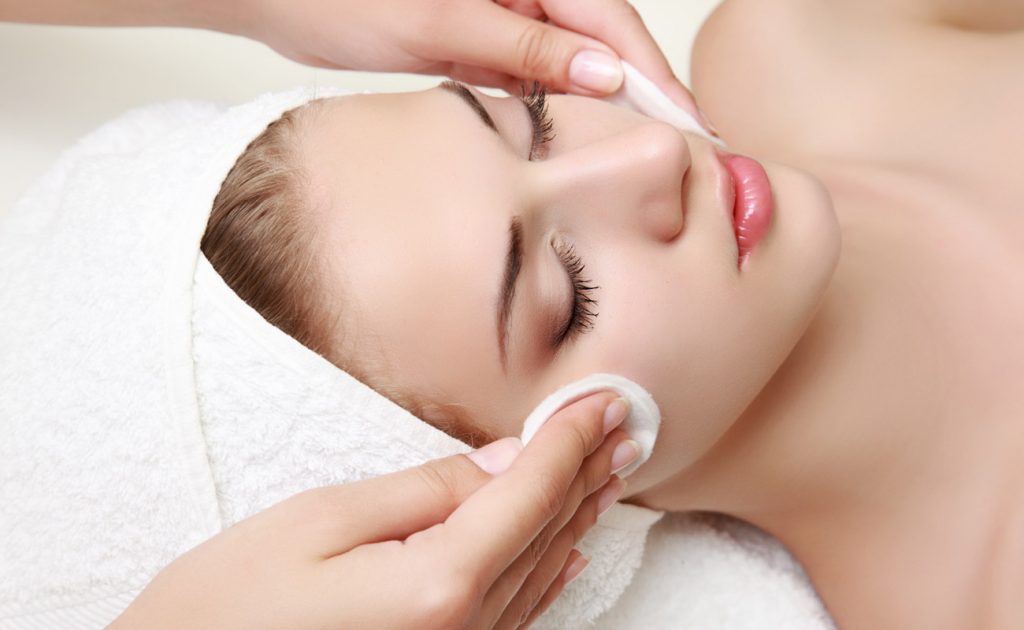 Close-up of a woman with eyes closed, receiving facial cleansing with cotton pads. She has a towel wrapped around her head.
