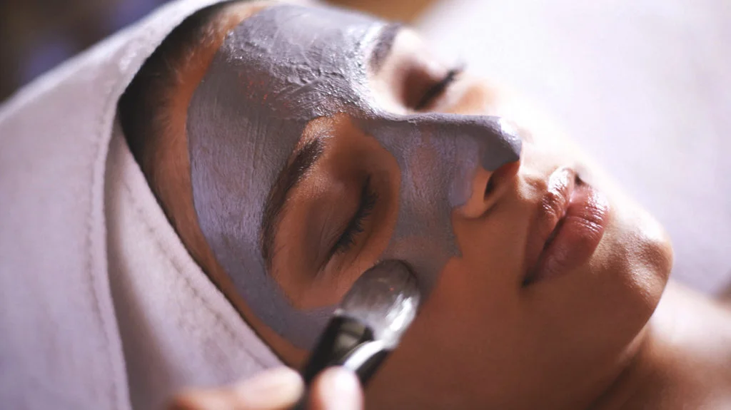 Close-up of woman with closed eyes receiving facial mask application with brush.